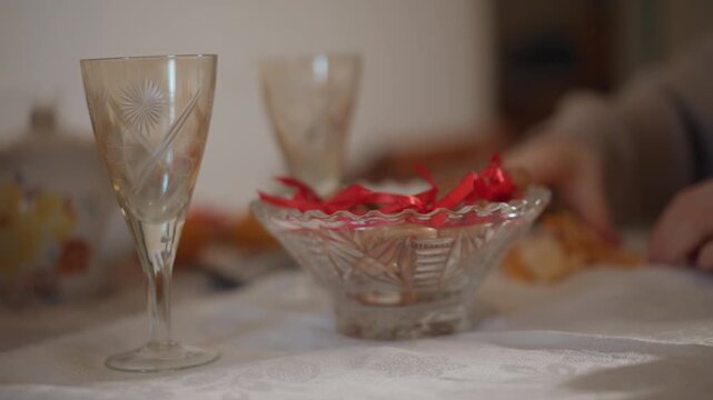 Caucasian woman placing bowl of wrapped red candy on linen cloth, crystal goblet beside, soft daylight, vintage porcelain teapot in background, subtle hospitality mood, refined detail shots