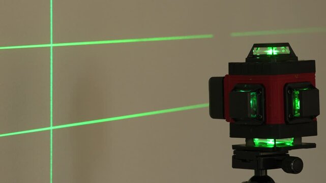 Red and black laser level mounted on a tripod projects bright green grid lines across a beige wall. Glowing green indicators light the device's surface