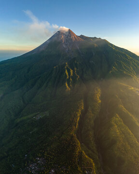 Aerial view of Mount Merapi with volcanic smoke rising from the crater, lush green forested slopes, and golden morning light in Cangkringan, Special Region of Yogyakarta, Indonesia.