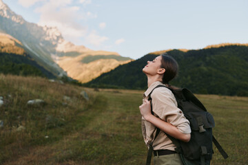 Obraz premium Young woman with a backpack hikes through a grassy valley beneath dramatic mountain scenery, looking upward with a calm, inspired expression in bright daylight.