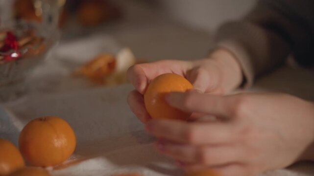 Hands peeling orange on table, soft warm light, slow domestic ritual with several whole oranges, linen surface and glass bowl blurred in background, close intimate framing showing skin texture