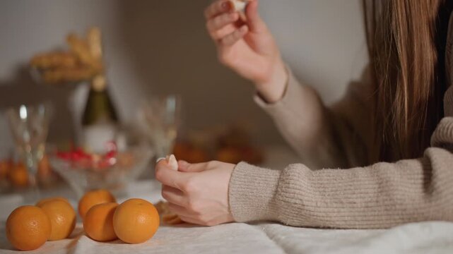 Caucasian woman peeling orange with hands, mandarin segments on table, glassware and bowl in background, cozy sweater, warm ambient light, intimate snack preparation for small gathering