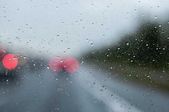 Close-up view of raindrop on glass windshield with blurred red taillight creating bokeh effect on gray rainy day