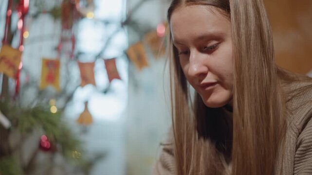Teen girl writing heartfelt holiday note at decorated table, warm window light, blurred tree and bunting, focused expression, pen on paper, envelope and ornaments nearby, cozy sweater, closeup