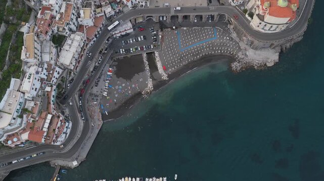 Aerial view of the coastal town of Atrani with its beach, rows of umbrellas, small boats moored in the clear water, and winding roads along the cliffs in Atrani, Campania, Italy.