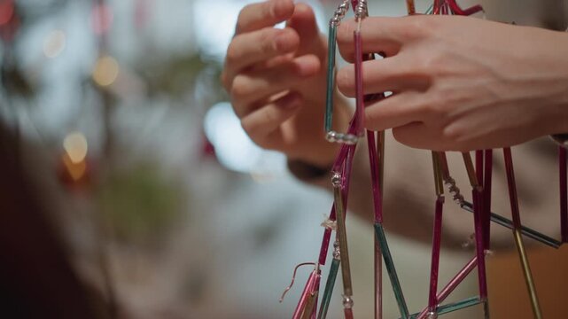 Closeup hands holding metallic garland caucasian woman carefully untangling colorful tinsel ornaments near blurred holiday lights, sweater sleeve visible, slow deliberate motions, warm indoor glow,