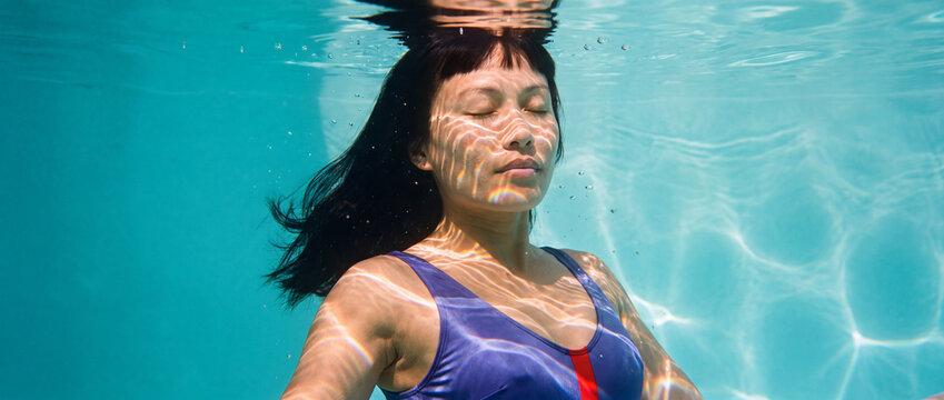 Woman underwater swimming with eyes closed in a clear blue pool, featuring bright sunlight reflections and floating dark hair, ideal for wellness, freedom, and summer themes