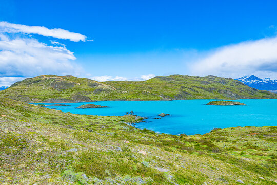 Brilliant azure blue waters of Lake Nordenskj&ouml;ld lapping against green grassy hills beneath cloudy skies in Torres del Paine, Chile.