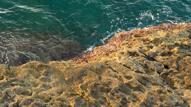 Dark green sea waves splashing against orange rocky coastline with textured volcanic looking rocks in bright sunlight