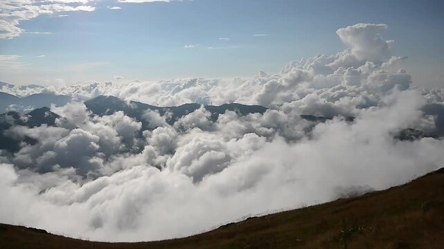 Sea of clouds rolls past mountain ridges with cumulus clouds above dry grassy slopes.