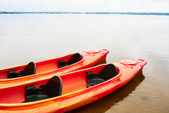 Commercial rental canoes resting on the sand by a calm lake. Empty tandem boats prepared for customers. Bright red paddling equipment on a river bank during a quiet overcast summer day in nature.