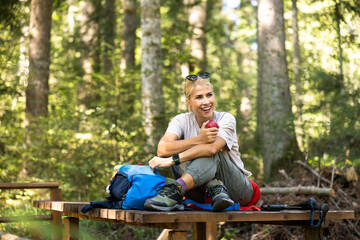 Fototapeta premium Woman hiker relaxing on a wooden bench, eating a fresh apple and smiling, taking a break in a green forest