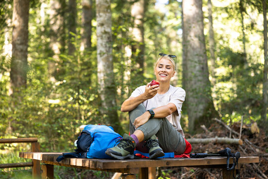 Young woman on a forest hike sitting on a wooden bench, smiling while eating a fresh red apple, pausing for a break