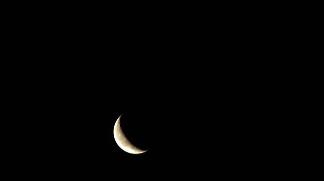 Time lapse of crescent moon rising against deep black cloudless night sky above horizon.