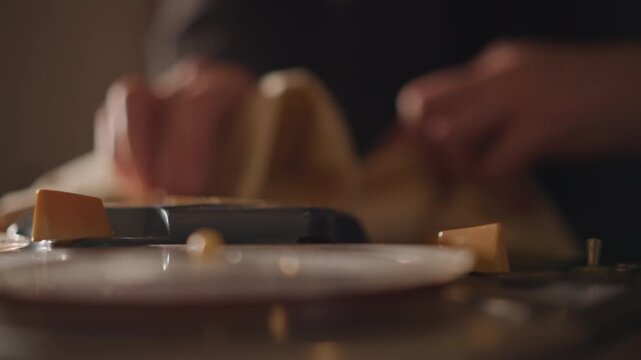 Closeup caucasian hands cleaning projector platter with cloth, warm cinematic lighting, dust removal, careful restoration gesture, tactile vintage equipment, moody lowlight atmosphere