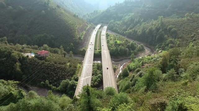 An aerial view shows road bridges and highway tunnels crossing a forested mountain valley landscape.