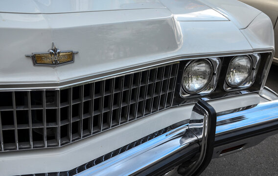 Close up of a white classic Chevrolet Caprice front end, featuring its egg crate chrome grille, dual round headlights, a chrome bumper, and a gold hood emblem