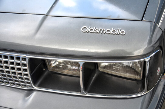 Close up of a grey vintage Oldsmobile front end, featuring dual recessed rectangular headlights, a chrome grid grille, and a silver script Oldsmobile logo on the hood