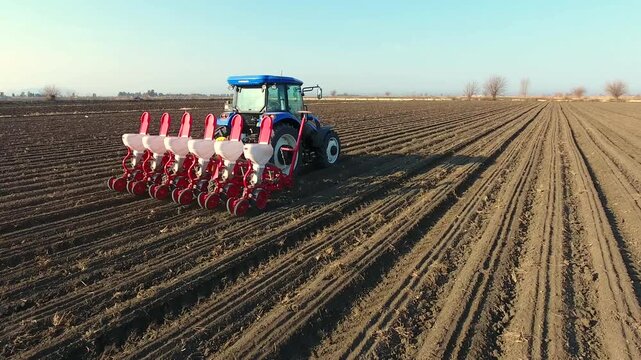 Aerial view of blue tractor planting fertilizer and seeds across wide brown field farmland.