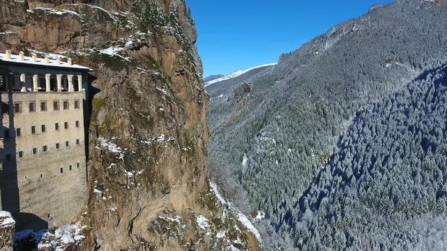 Aerial drone view captures snow covered Sumela Monastery clinging to steep rocky mountain cliff.