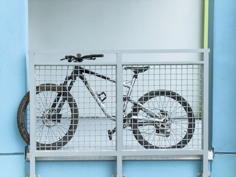 Eye-level view of a multi-speed bicycle parked on an otherwise empty apartment balcony, with its front wheel protruding a little from a mesh guardrail. Motifs of transportation, storage, and security.