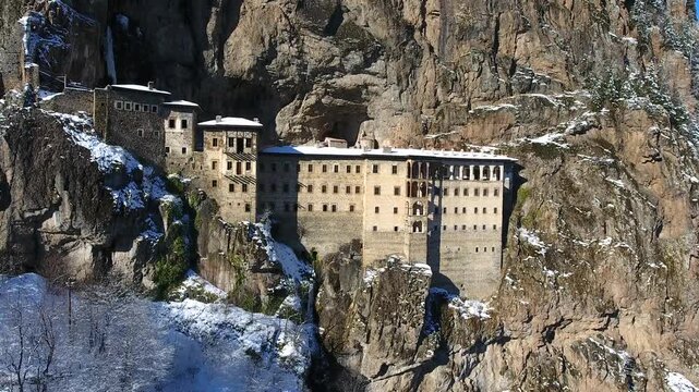 Aerial drone view captures snow covered Sumela Monastery clinging to steep rocky mountain cliff.