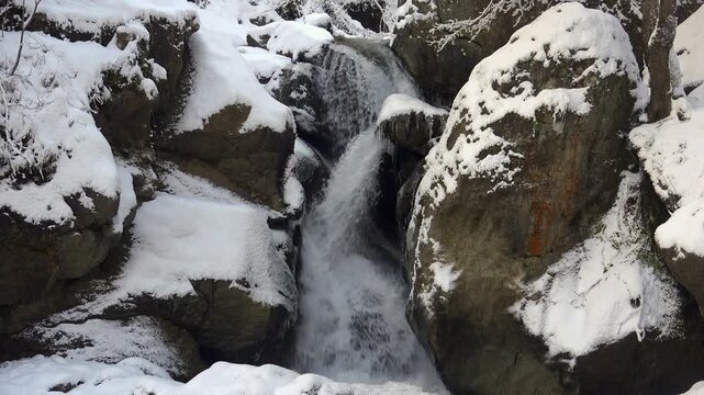 Small winter stream flows over snowy rocky slope with icy water cutting through stones.