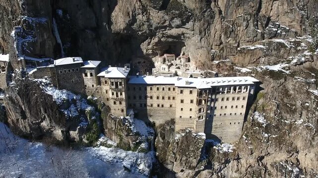 Aerial drone view captures snow covered Sumela Monastery clinging to steep rocky mountain cliff.