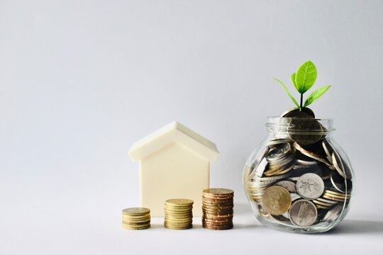 Concept of saving for a home showing ascending columns of coins, a miniature house model, and a glass coin jar topped with a growing green leaf bud on a white background.