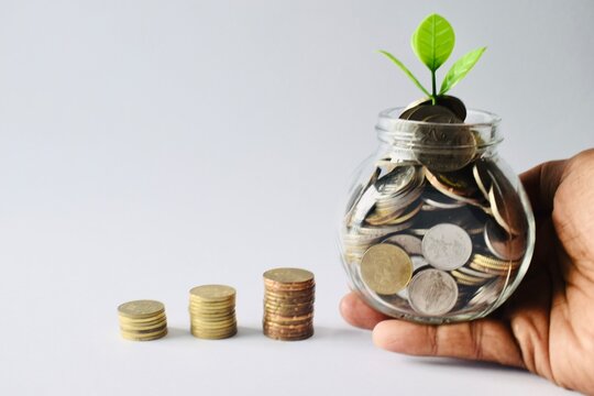 A hand holding a glass savings jar filled with coins and a green plant shoot growing out of the top, positioned next to three growing stacks of coins on a plain white background.
