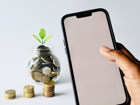 Close-up of a person holding a smartphone with a blank mockup screen against a white background, with a savings glass jar filled with coins and a growing green plant in the blurred background.