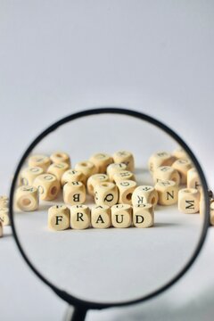 A magnifying glass focused on wooden letter blocks that spell the word FRAUD, surrounded by a scattered collection of alphabet beads on a bright background.