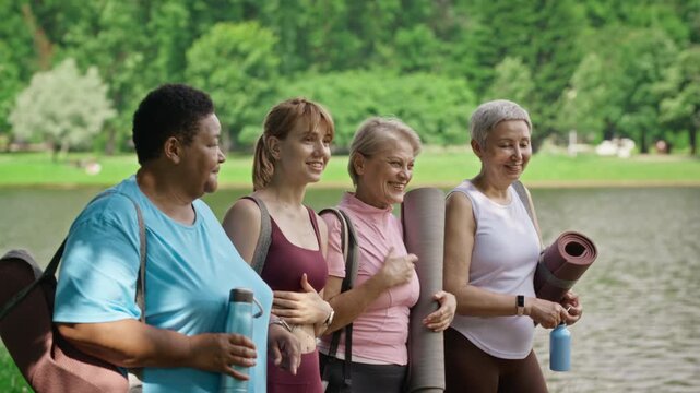 Diverse multiethnic women, including senior and young adult trainer, holding yoga mats and talking near lake during tracking shot. Footage is ideal for wellness marketing campaigns