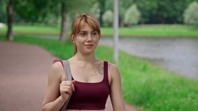 Young adult Caucasian woman carrying yoga mat walking along park path near lake, tracking shot. Clip is ideal for promoting outdoor fitness, mental wellbeing, and healthy lifestyle choices