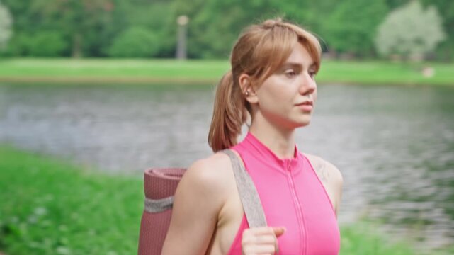 Young adult Caucasian woman walking along park path with yoga mat on shoulder, tracking shot. Video is perfect for commercial use in fitness, wellness, and healthy lifestyle campaigns