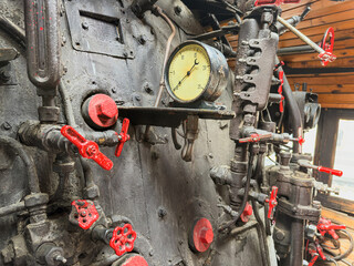 Steam locomotive cab control panel, intricate machinery details, red valves, circular pressure gauge, dark metallic surface, wooden ceiling glimpse, indoor museum workshop setting, even bright © Mister