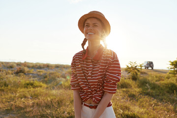 Obraz premium Smiling young woman in a straw hat and striped shirt stands in a field at sunset, embodying a joyful lifestyle theme with warm sunlight casting a soft glow on her face outdoors.