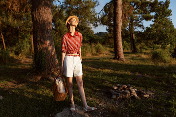 Obraz premium A redheaded woman lifestyle enjoying golden hour in a forest park. Wearing straw hat and summer clothes holding a picnic basket. Concept for outdoor leisure, relaxation, and nature travel.