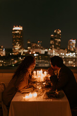 Fototapeta premium For their rooftop anniversary celebration, the couple enjoys city lights and candles while dining and laughing