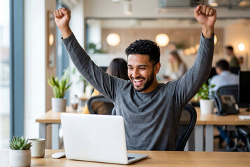 Fototapeta premium Happy young man celebrating success at his desk with raised fists in a busy modern office