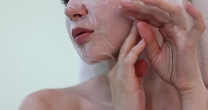 Young woman putting sheet mask onto her face on light grey background, closeup