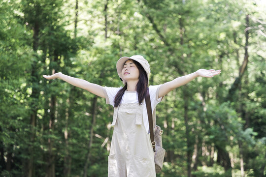 A young woman stands with outstretched arms, eyes closed, enjoying fresh air in a dense green forest. She wears a hat, overalls and carries a backpack, surrounded by lush trees.