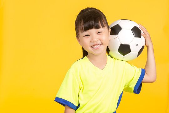 The smiling young girl leans slightly to the side on the yellow background, balancing the soccer ball casually behind her head with one hand, looking up playfully with a joyful expression in her eyes.
