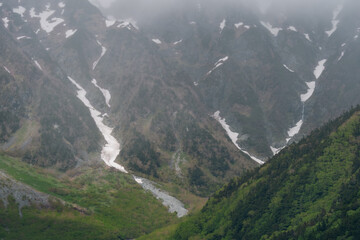 Obraz premium Mountain valley snow forest alpine peak mist cloud rocky slope green hill river landscape with rugged terrain, foggy sky, glacier patch, wilderness, and remote nature scenery