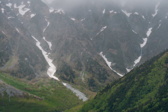 Mountain valley snow forest alpine peak mist cloud rocky slope green hill river landscape with rugged terrain, foggy sky, glacier patch, wilderness, and remote nature scenery