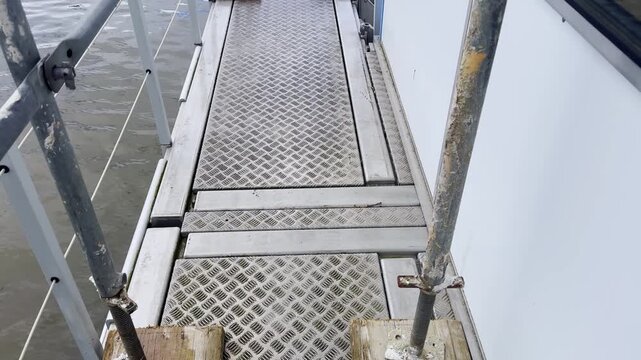 Narrow floating platform walkway beside calm lake water, with metal grating floor, scaffold poles, railings, wooden base plates and temporary event structure details.