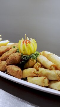 Close-up of a hand using metal tongs to pick up a crispy spring roll from a large platter of fried appetizers and kibbeh indoors, perfect for catering and restaurant promotions.