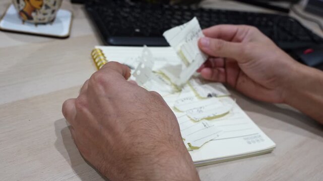Close-up of a hand tearing handwritten notes from a spiral notebook on an office desk, with a keyboard, pen, and decorative mug in the background.
