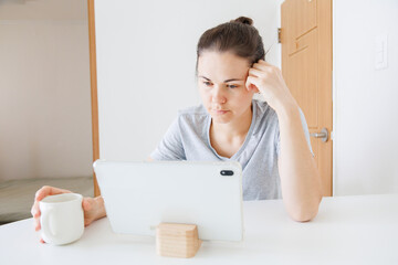Woman focused on a tablet at home while holding a cup of coffee. Online activity, internet browsing, digital technology, tablet use and home lifestyle. © soya