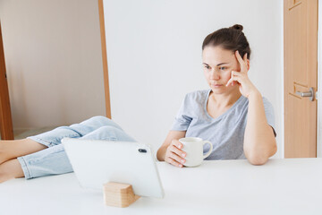 Woman relaxing at home with feet on the table, drinking coffee and using a tablet. Casual home lifestyle, digital entertainment, leisure time and mobile technology. © soya